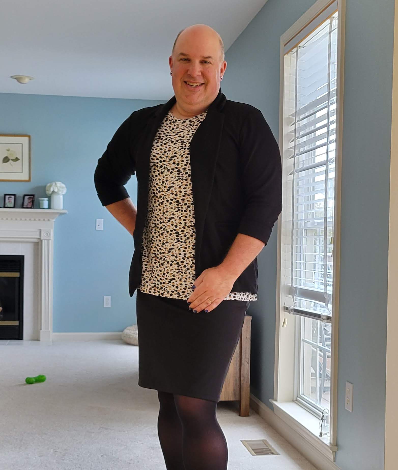 a man stands in a living room. he's wearing a leopard print top and black skirt, with a black blazer over it.
