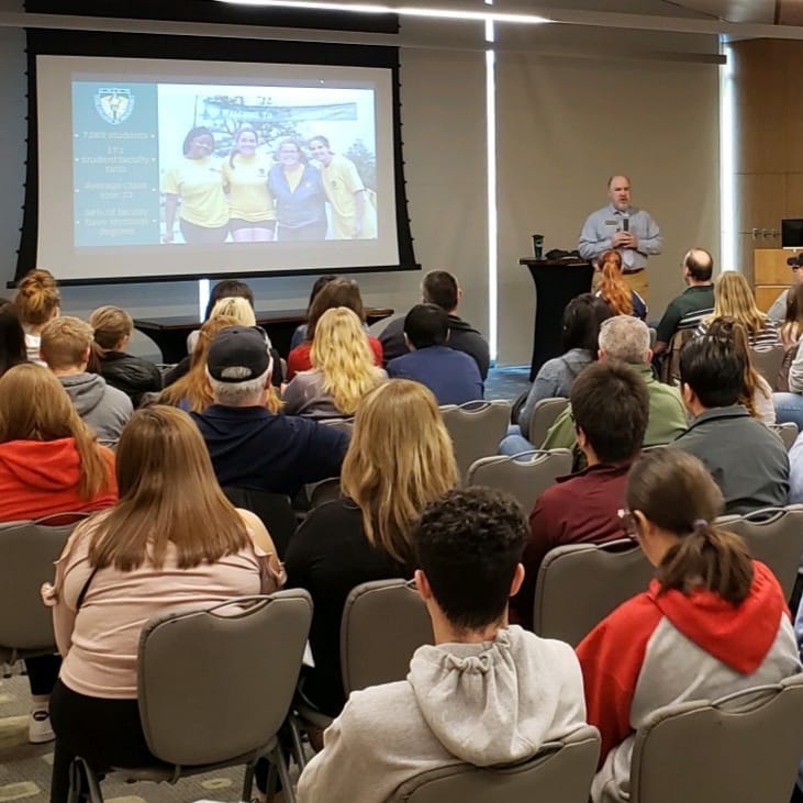 dozens of students and families face away from the camera towards a screen on the back wall; a man is shown with a microphone next to the screen, presenting.