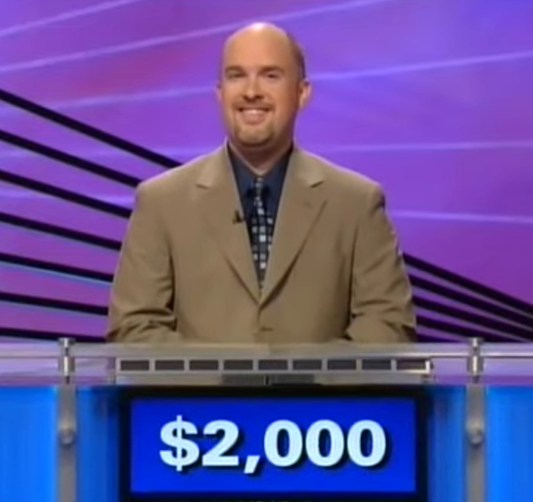 title a man in a tan suit stands behind a game show podium. His score is listed on the front of $2,000.